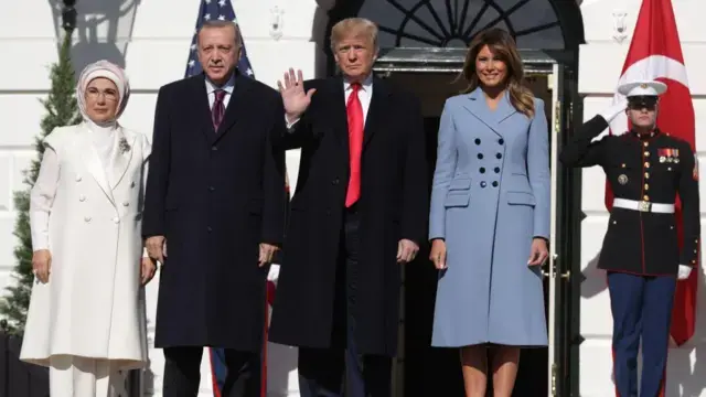 Emine Erdoğan, Recep Tayyip Erdoğan, Donald Trump ve Melania Trump 2019'da Beyaz Saray kapısında poz veriyor. Emine Erdoğan beyaz, Melania Trump mavi uzun kaban giymiş. Güneşli bir gün. Erdoğan ve Trump da siyah kaban giyiyor. İçlerinde takım elbise var. 